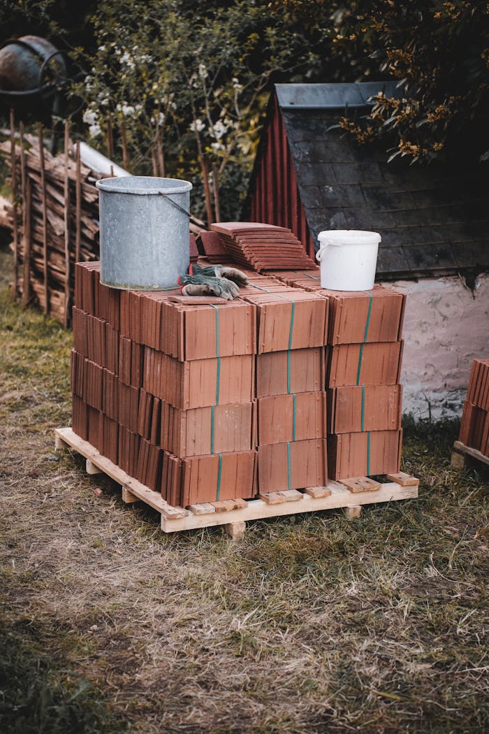 Pallet of stacked roof tiles with metal and plastic buckets outside near a shed.